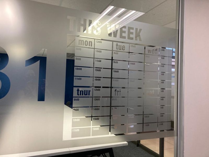 A modern office space featuring a frosted glass partition with a printed weekly schedule, providing a professional and organized work environment.