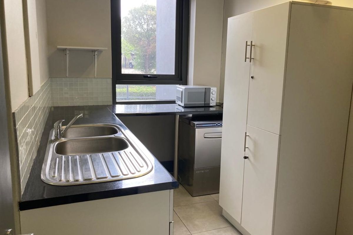 A functional office kitchenette featuring a stainless steel double sink, tiled backsplash, laminate countertops, and built-in storage cabinetry.