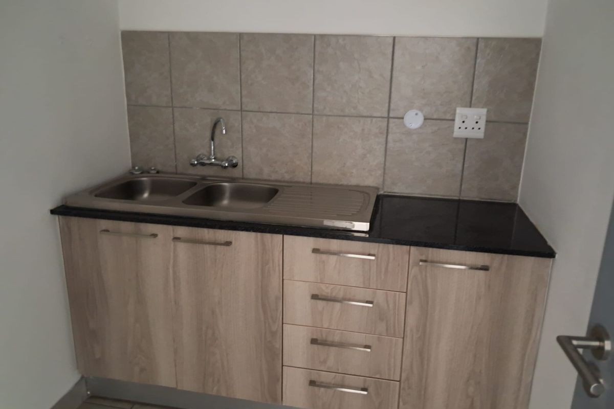 A compact kitchenette featuring light wood-grain cabinetry, a dark granite countertop, a stainless steel double sink, and neutral tiled splashback.