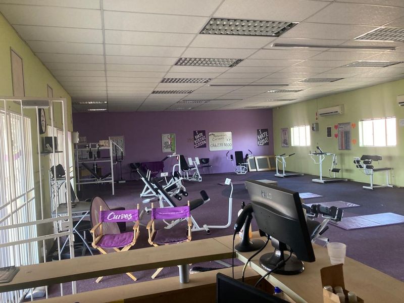 A commercial gym space featuring a reception area, suspended ceiling with recessed lighting, wall-mounted air conditioning, and open floor plan for fitness equipment.