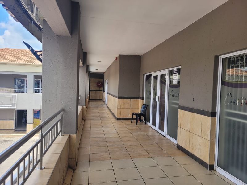 A covered outdoor walkway in a retail complex featuring tiled flooring, metal railings, and glass storefront entrances.