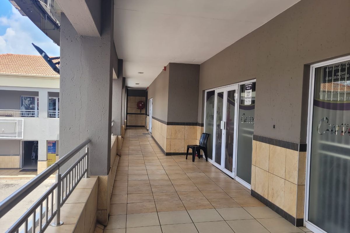 A covered outdoor walkway in a retail complex featuring tiled flooring, metal railings, and glass storefront entrances.