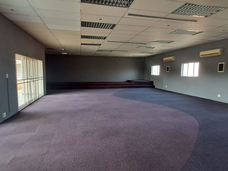 Spacious open-plan retail or commercial area featuring a raised stage, suspended ceiling, wall-mounted air conditioning units, and large windows with security bars.