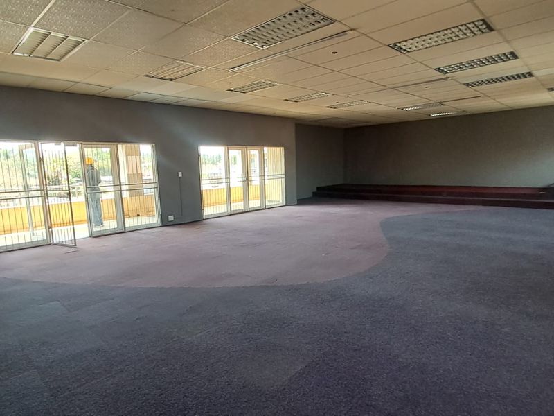 Large open-plan retail or commercial space featuring a suspended ceiling, recessed lighting, carpeted flooring, and a raised platform area with access to a balcony.