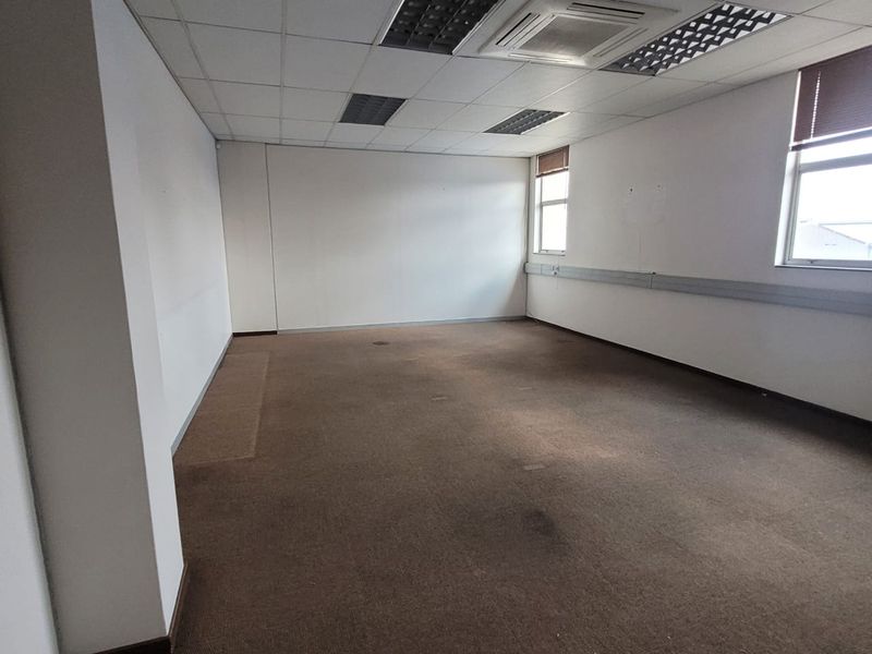 An empty, open-plan office space featuring a suspended ceiling, recessed lighting, and carpeted flooring with wall-mounted trunking.