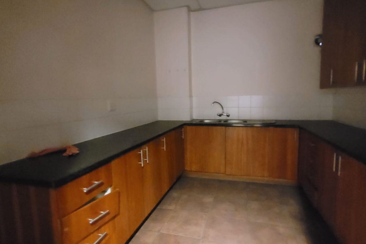 A functional office kitchenette featuring wooden cabinetry, dark countertops, a double sink, and tiled flooring. The space is currently vacant.