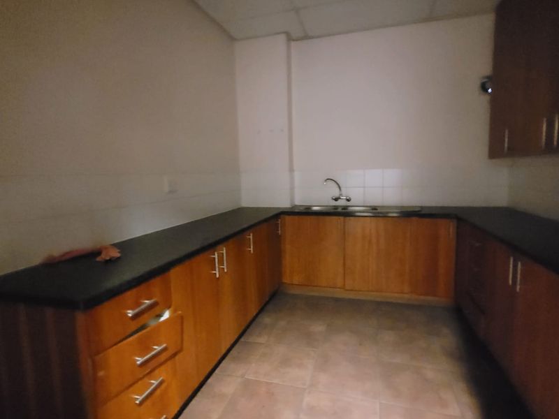 A functional office kitchenette featuring wooden cabinetry, dark countertops, a double sink, and tiled flooring. The space is currently vacant.