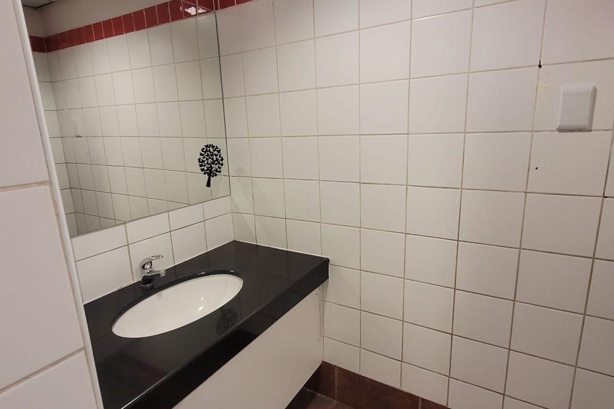 A functional office bathroom featuring a vanity basin with a dark countertop, wall-mounted mirror, and white tiled walls with a red accent border.