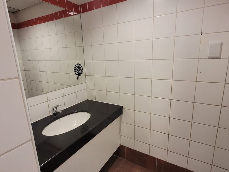 A functional office bathroom featuring a vanity basin with a dark countertop, wall-mounted mirror, and white tiled walls with a red accent border.