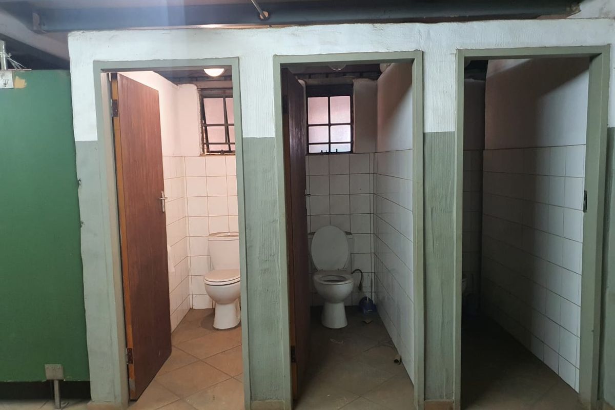 A row of three industrial-style toilet cubicles featuring tiled walls, ceramic toilets, and exposed ceiling beams.