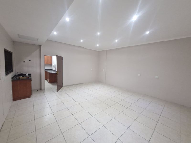 A spacious, empty office area featuring neutral tiled flooring, recessed lighting, and a small integrated kitchenette with granite countertops.