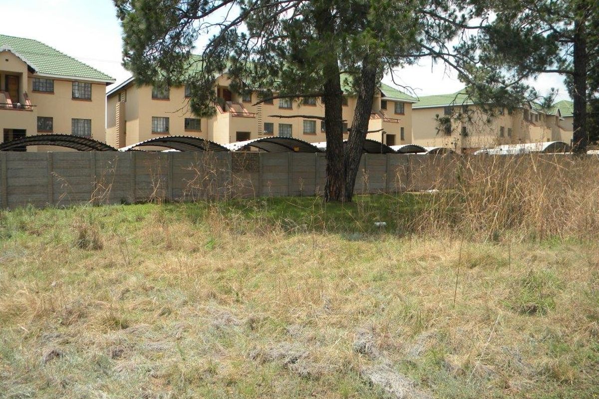 A view of an undeveloped residential plot featuring dry grass, mature trees, and a precast concrete boundary wall separating it from neighboring apartment complexes.
