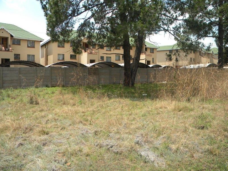 A view of an undeveloped residential plot featuring dry grass, mature trees, and a precast concrete boundary wall separating it from neighboring apartment complexes.