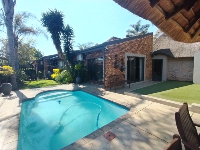 A well-maintained outdoor pool area featuring a brick-clad home exterior, a thatched roof section, and a paved patio space.