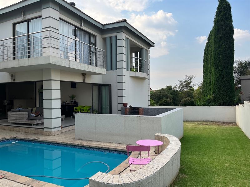 Rear view of a double-story house featuring a swimming pool, a built-in braai area, and an upper-level balcony overlooking the garden.