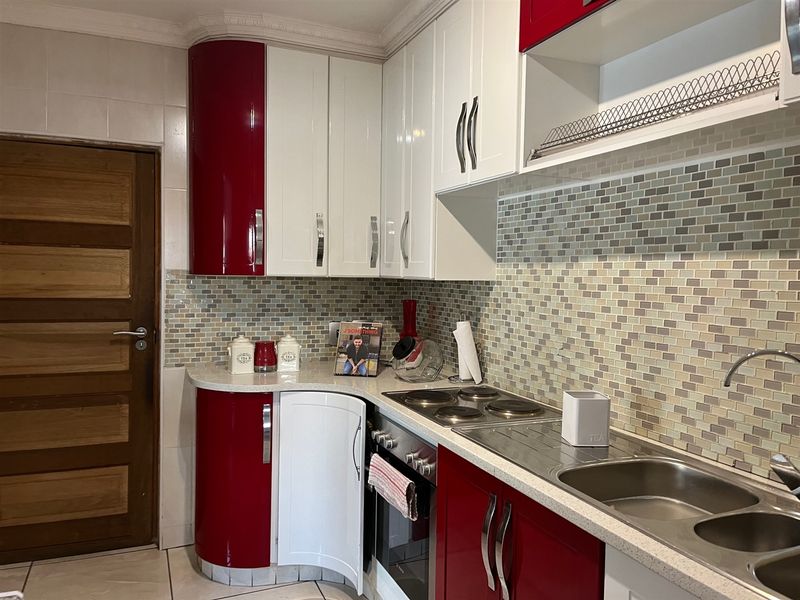 A compact kitchen featuring high-gloss red and white cabinetry, a mosaic tile backsplash, stone countertops, and integrated electric cooking appliances.