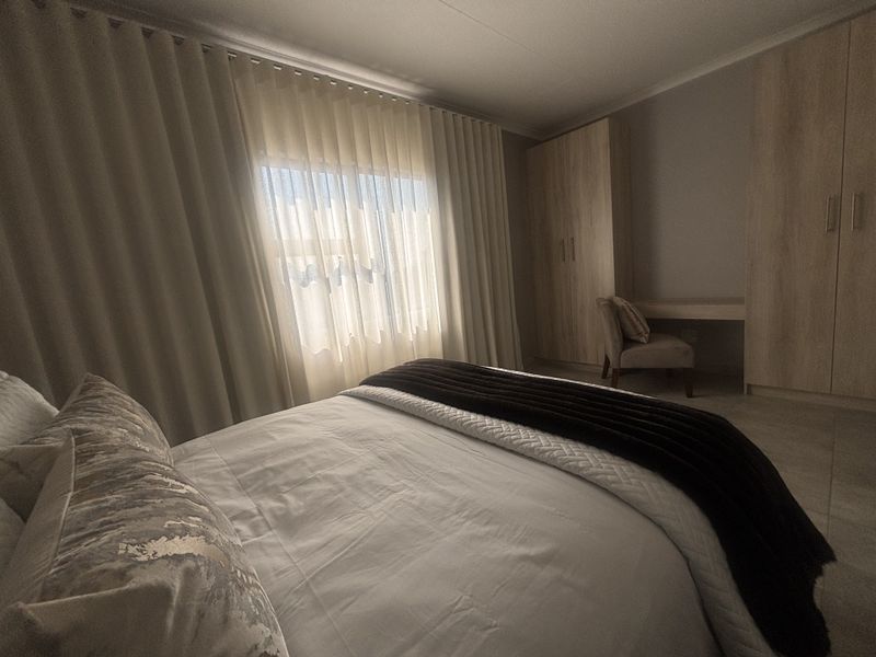 A well-maintained bedroom featuring built-in wooden cupboards, a matching integrated vanity, and neutral tiled flooring with natural light filtering through curtains.