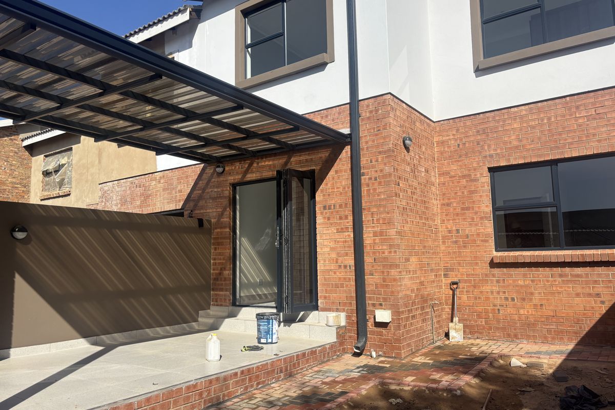 A rear exterior view featuring a covered patio area with modern black-framed folding doors, brick facade, and paved flooring.