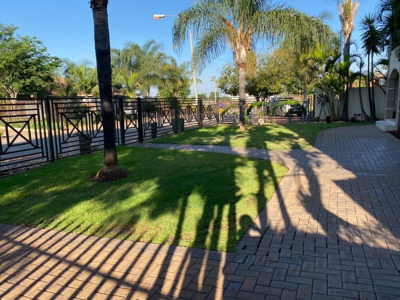 A well-maintained front garden area featuring a paved driveway, manicured lawn, and secure metal perimeter fencing.