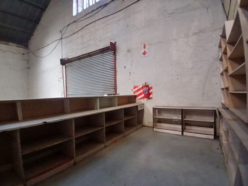 A vacant retail utility or storage space featuring built-in shelving, a roller shutter door, and exposed industrial roofing.