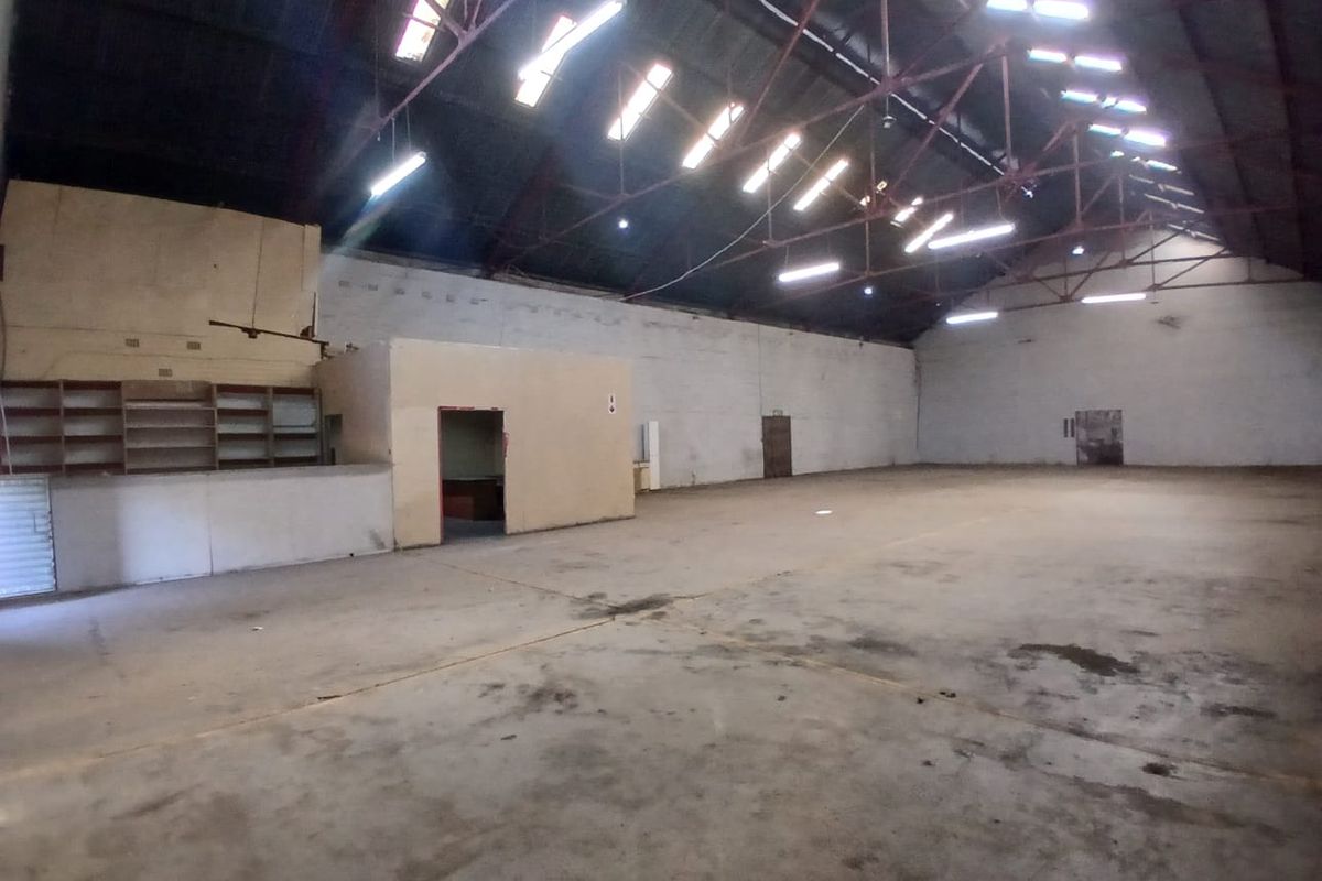 Spacious industrial utility space featuring high exposed steel roof trusses, skylights, concrete flooring, and a partitioned office area with built-in shelving.