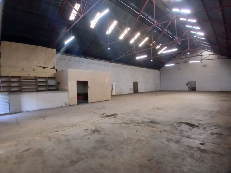 Spacious industrial utility space featuring high exposed steel roof trusses, skylights, concrete flooring, and a partitioned office area with built-in shelving.