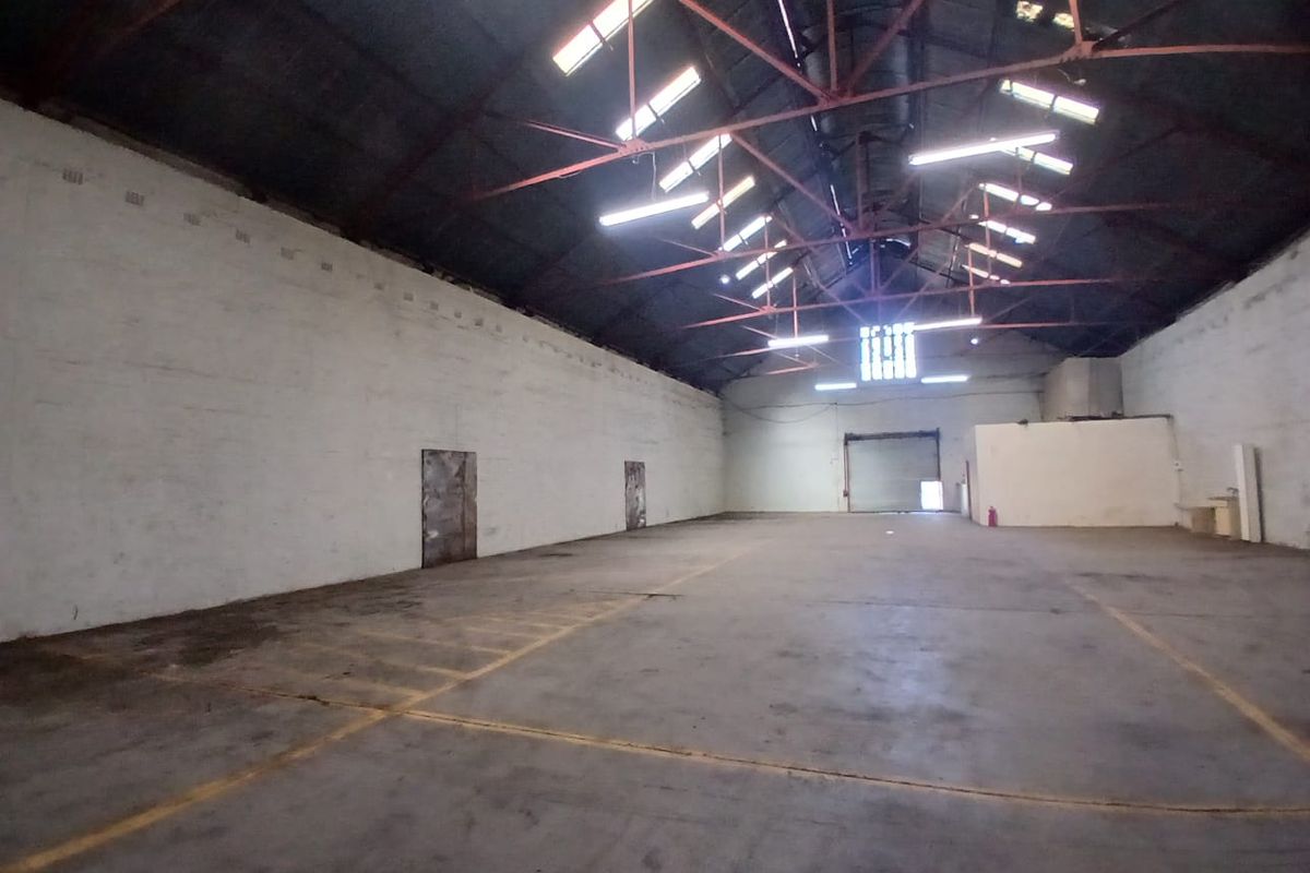 Spacious industrial warehouse unit featuring high exposed steel roof trusses, concrete flooring, skylights, and a large roller shutter door for easy access.