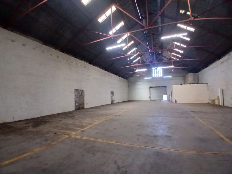 Spacious industrial warehouse unit featuring high exposed steel roof trusses, concrete flooring, skylights, and a large roller shutter door for easy access.