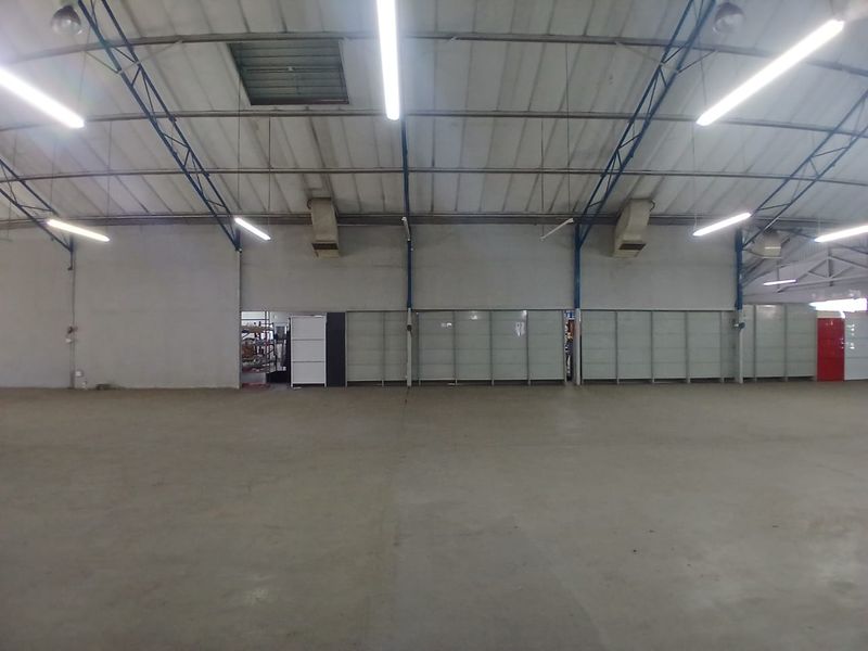 Spacious industrial warehouse area featuring high ceilings with exposed steel trusses, concrete flooring, and overhead fluorescent lighting.