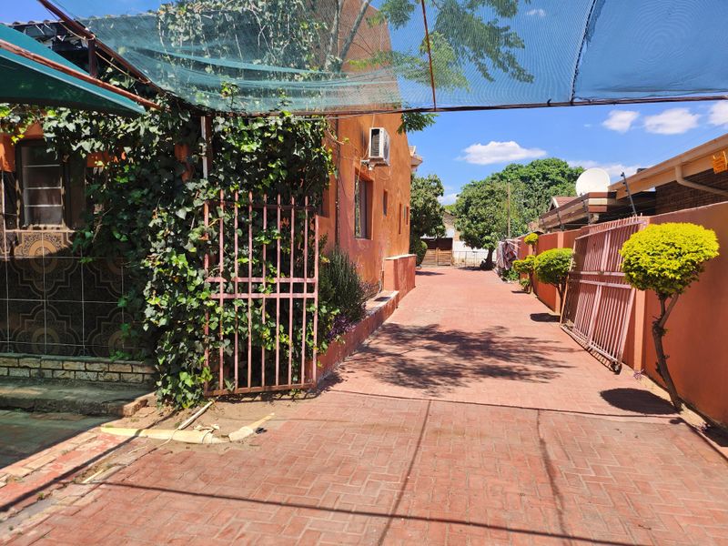 A paved driveway leading to the side of the house, featuring a metal security gate, perimeter walling, and overhead shade netting.