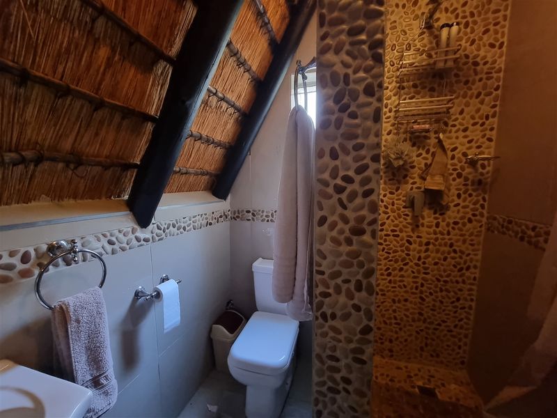 A unique bathroom featuring a rustic thatched roof, pebble-tiled shower enclosure, and standard sanitary fixtures in a compact layout.
