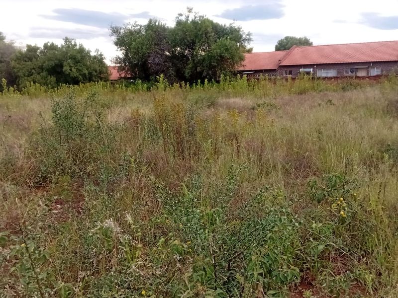 A view of a vacant residential plot featuring overgrown grass and shrubs, with neighboring properties visible in the background.