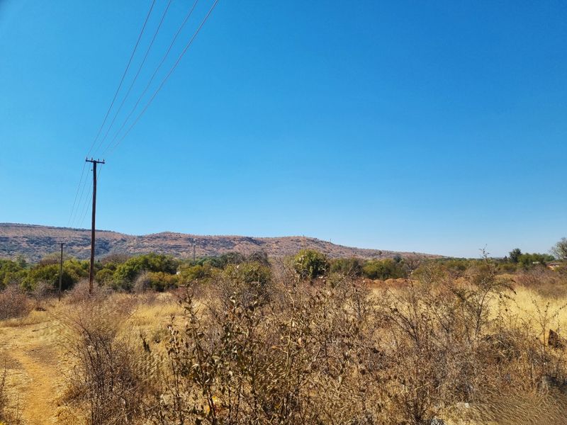 A view of the vacant residential land featuring natural vegetation, a utility pole, and distant mountain scenery under a clear blue sky.