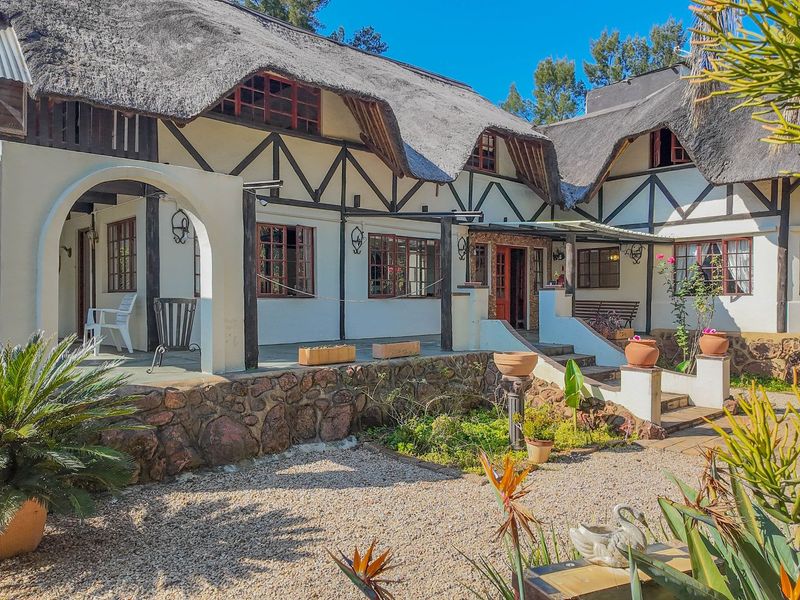 A charming rear elevation of a thatched-roof small holding featuring timber-framed windows, stone-clad base, and a spacious covered patio area.