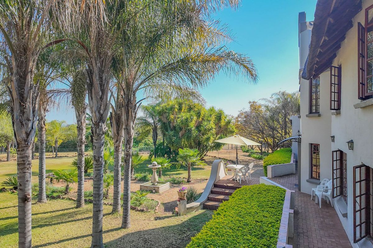 A spacious rear garden featuring a brick-paved patio, stone fountain, and mature palm trees, adjacent to a multi-story home with a traditional thatched roof.