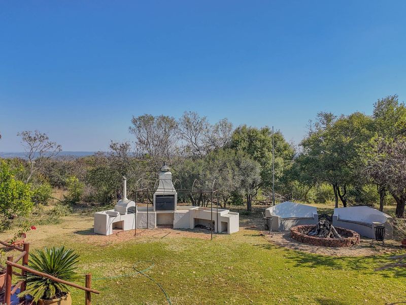 Spacious outdoor entertainment area featuring a built-in braai, pizza oven, stone fire pit, and masonry seating set within a landscaped garden.
