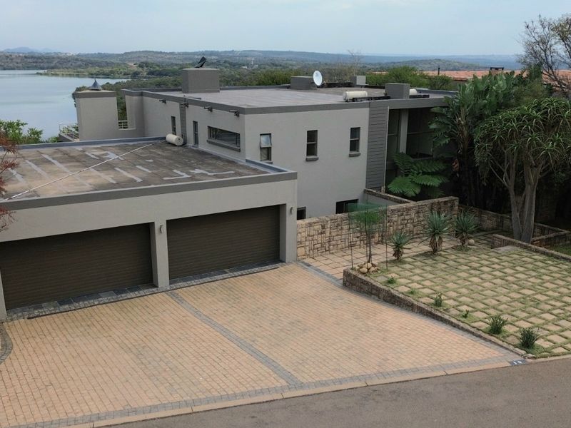 Aerial view of a modern multi-level house featuring a double garage, paved driveway, stone-clad exterior walls, and a landscaped garden overlooking a lake.