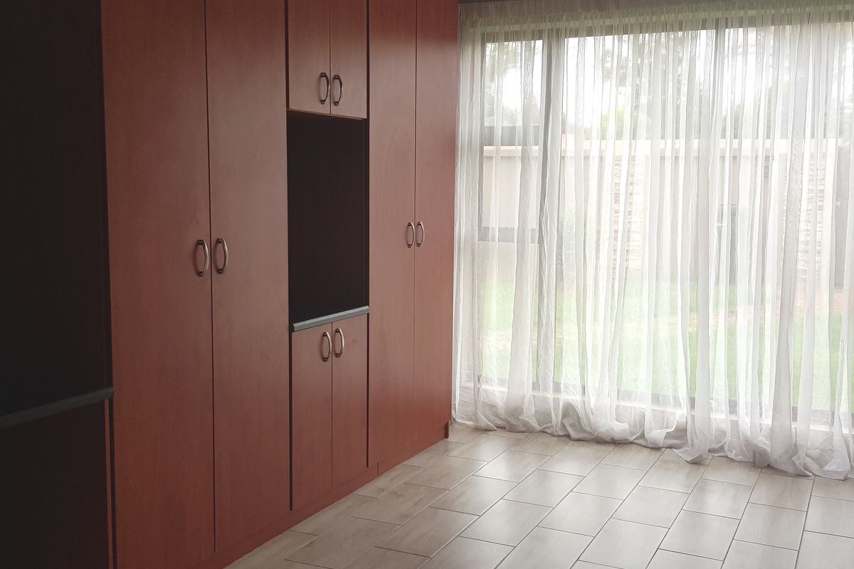 A bright, empty bedroom featuring built-in wooden cupboards, tiled flooring, and a large window with sheer curtains overlooking the garden.