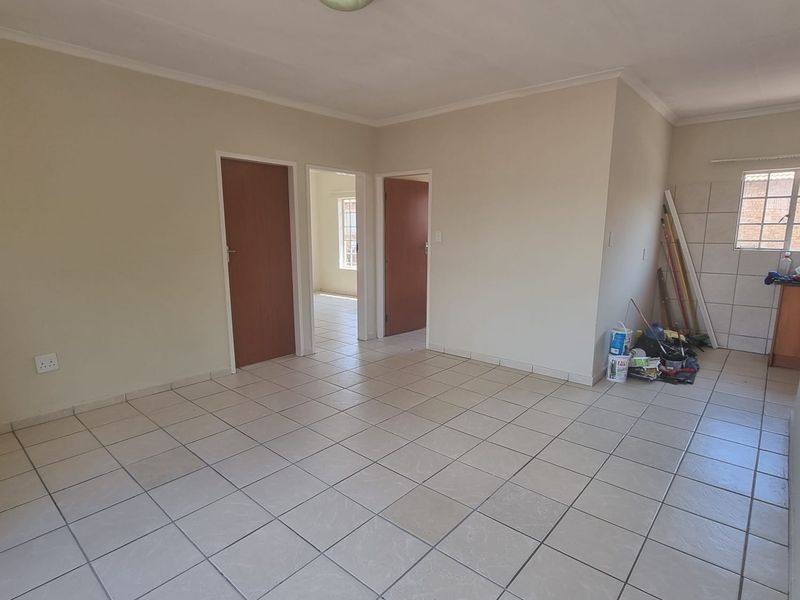 An open-plan living area featuring neutral ceramic floor tiles, multiple interior doorways, and a tiled kitchen splashback area in a vacant, well-lit space.