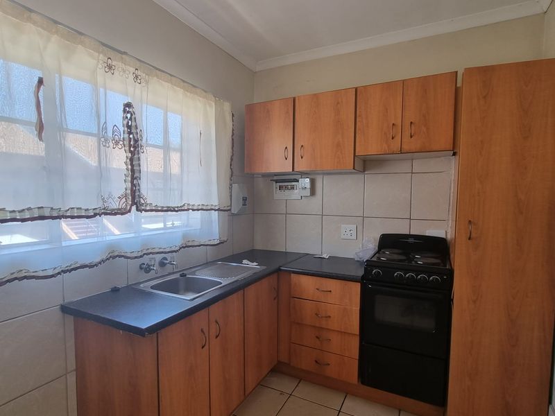 A compact kitchen featuring laminate cabinetry, tiled backsplash, stainless steel sink, and a freestanding stove, with some visible wear to the floor tiles.