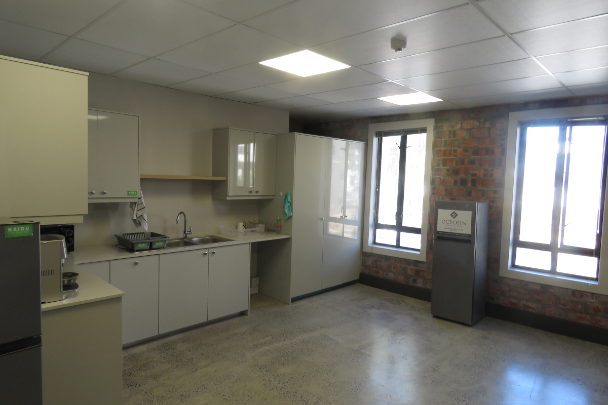 A functional office kitchenette featuring modern cabinetry, a stainless steel sink, and exposed brick wall accents with polished concrete flooring.