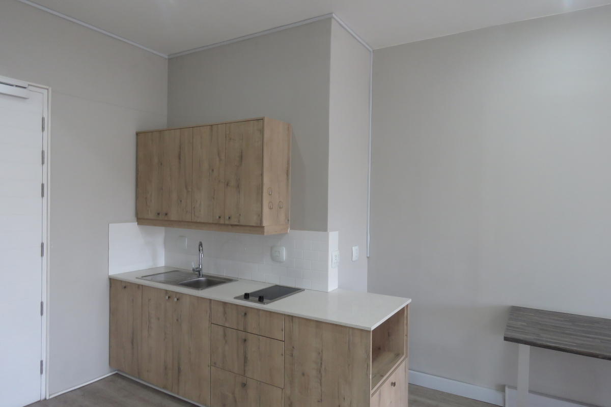 A compact kitchenette featuring light wood-grain cabinetry, a stainless steel sink, a built-in hob, and white subway tile backsplash in a modern office space.