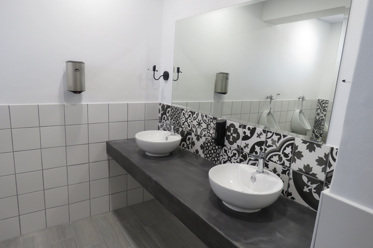 Modern office restroom featuring a dual-basin vanity with concrete-style countertop, decorative patterned tiling, and wall-mounted fixtures.