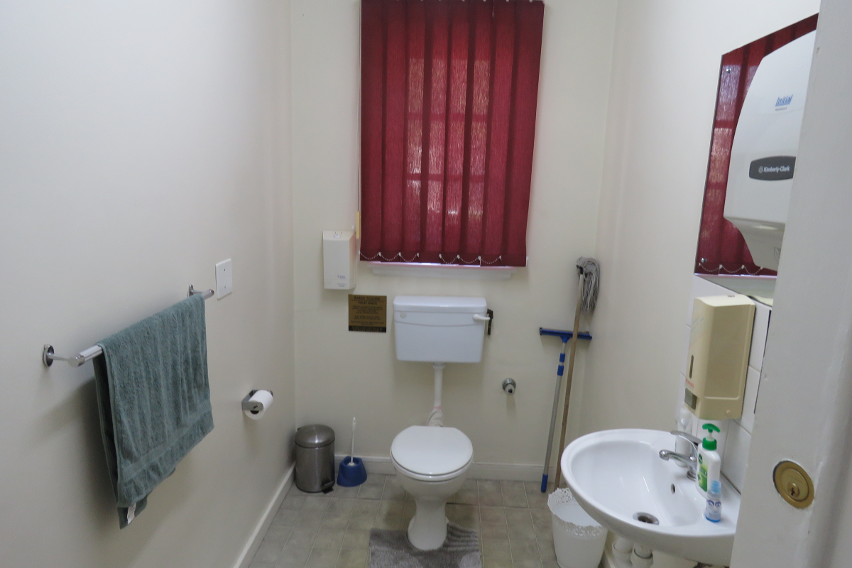A functional office restroom featuring a standard toilet, wall-mounted basin, and vertical window blinds.