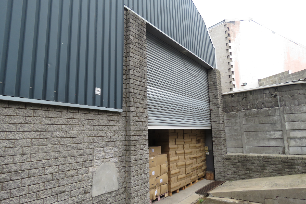 Industrial warehouse exterior featuring a large roller shutter door, corrugated metal cladding, and a concrete access ramp with perimeter security fencing.