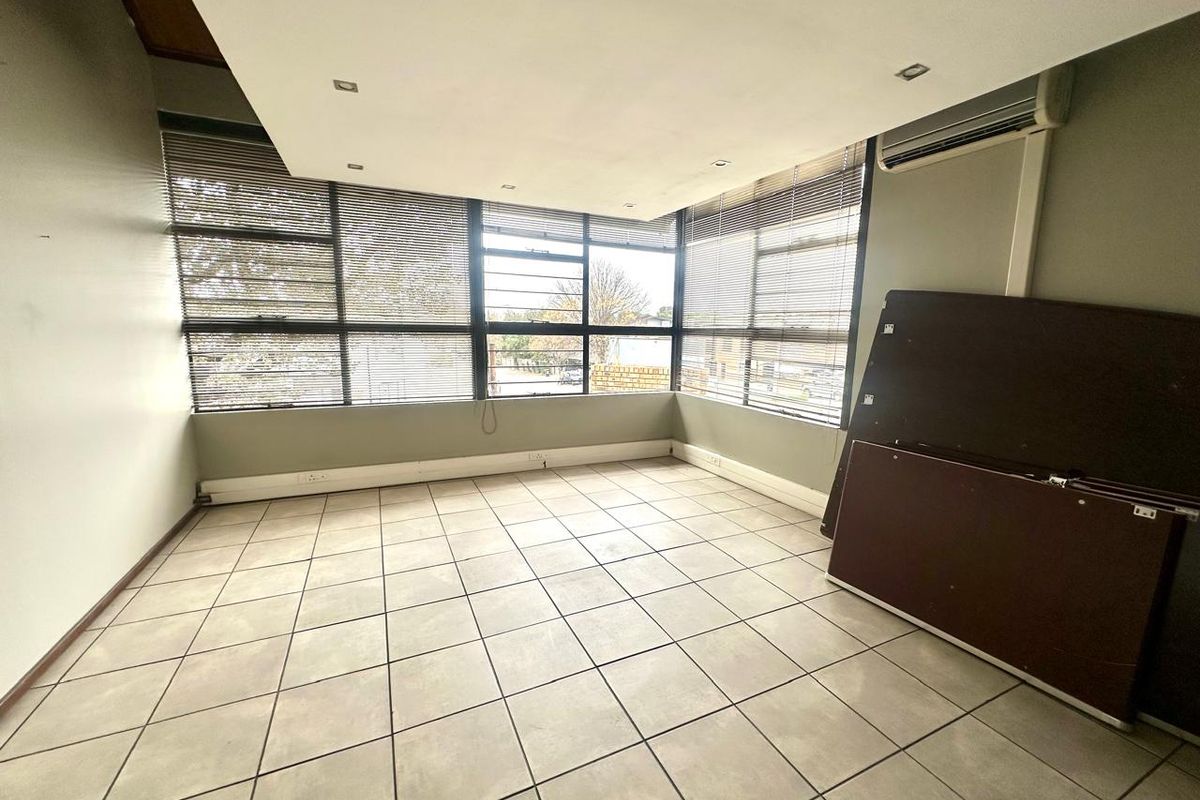 A bright, open-plan office space featuring large windows with fitted blinds, tiled flooring, and a wall-mounted air conditioning unit.