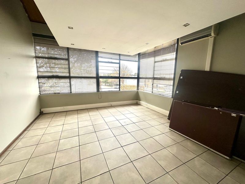 A bright, open-plan office space featuring large windows with fitted blinds, tiled flooring, and a wall-mounted air conditioning unit.