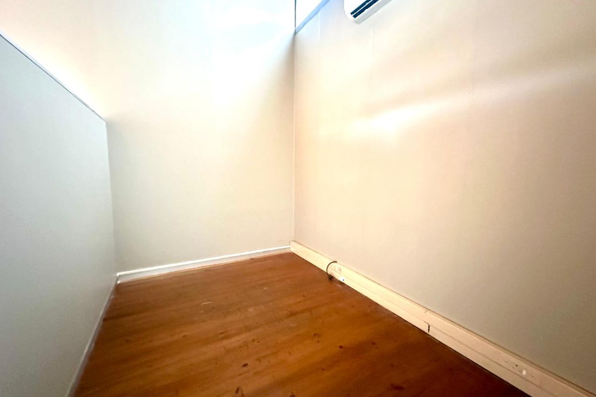 A compact, empty office space featuring timber flooring, a wall-mounted air conditioning unit, and neutral wall finishes.