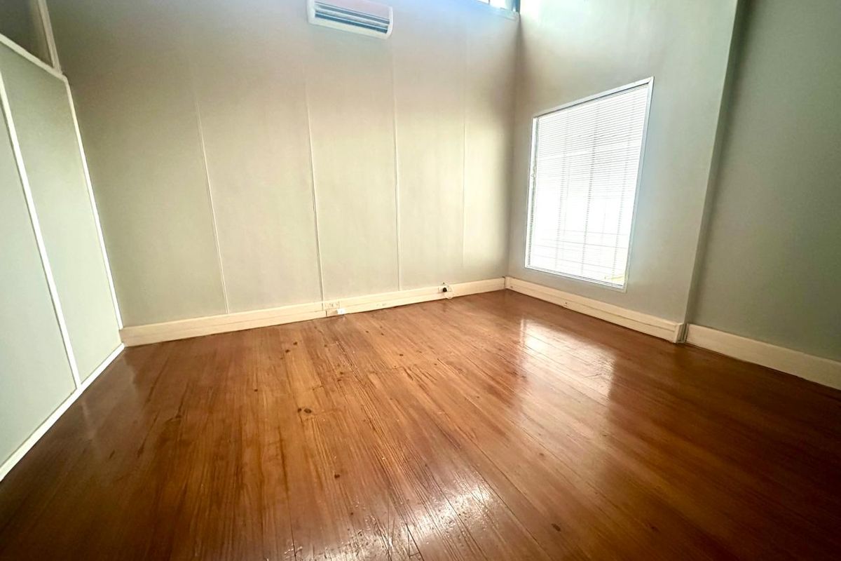 A bright, empty office space featuring polished timber flooring, high ceilings, a wall-mounted air conditioning unit, and large windows with fitted blinds.