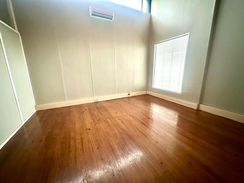 A bright, empty office space featuring polished timber flooring, high ceilings, a wall-mounted air conditioning unit, and large windows with fitted blinds.